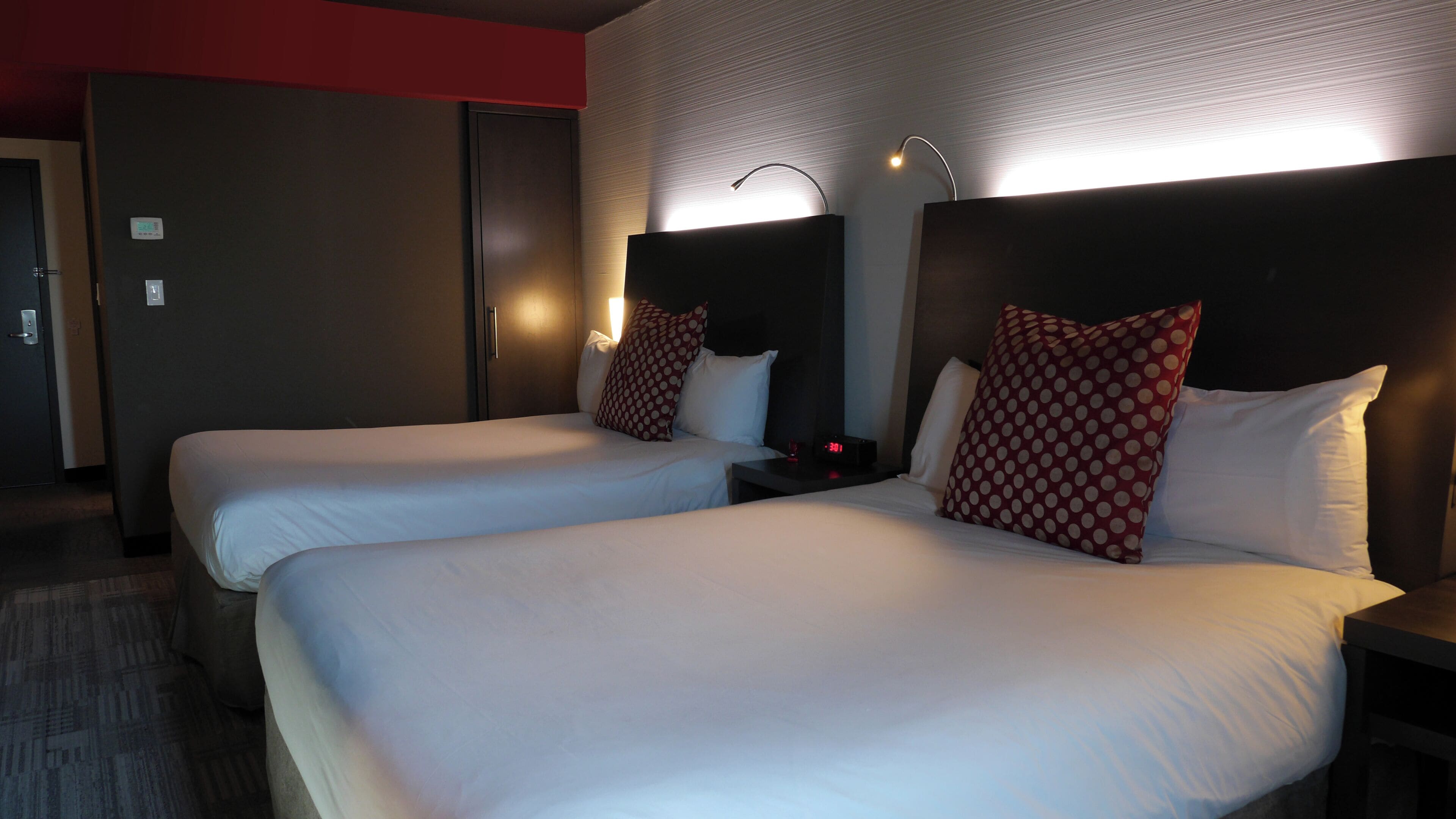 A hotel room with two beds, each with white bedding and red polka dot pillows. There is a nightstand with a clock and a lamp between the beds. The room has a red accent wall and a door to the left.