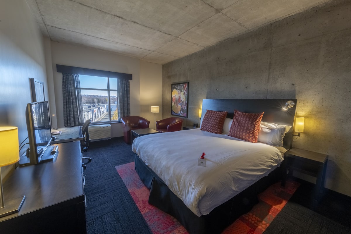 Habitación de hotel con una cama grande, un escritorio con lámpara, una silla, una lámpara de escritorio, una ventana con cortinas y un cuadro en la pared.