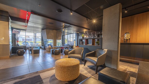 A modern lobby with seating areas, a large window offering a view of parked cars, and a bar area with a plant and decorative items. - Grand Times Hotel Quebec City Airport (L'Ancienne-Lorette)
