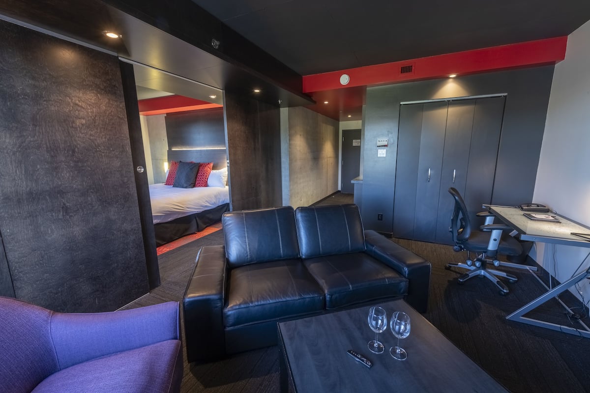 Una habitación de hotel moderna con un sofá de cuero, una mesita con vasos, un escritorio con una silla y una cama con almohadas rojas.