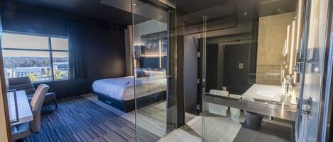 Habitación de hotel moderna con una cama grande, un escritorio, ducha envuelta en vidrio y vistas a la ciudad.