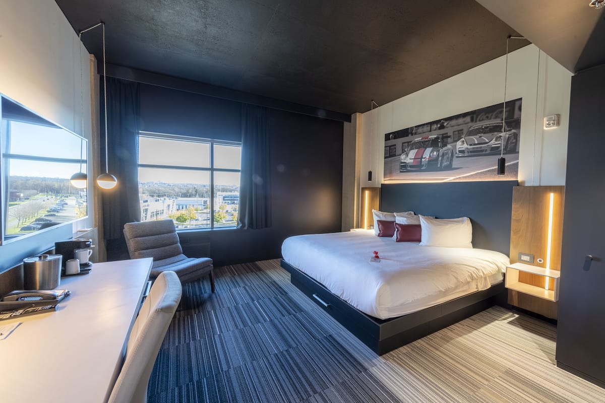 Una habitación de hotel moderna con una cama grande, un escritorio con una silla, vistas de la ciudad por la ventana y una obra de arte colgada en la pared que representa autos de carrera.