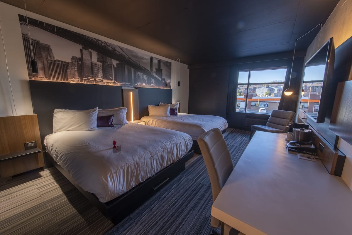 Habitación de hotel con dos camas, un escritorio y un amplio ventanal con vistas a la ciudad.