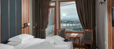 Habitación de hotel con una cama grande, una silla, una mesita y vistas a un paisaje urbano a través de la ventana.