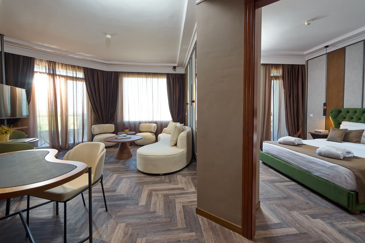 A hotel room with a green upholstered bed, a round coffee table, two armchairs, a sofa, and a television.