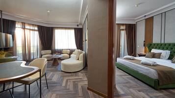 Habitación de hotel con una cama tapizada de verde, una mesa de centro redonda, dos sillones, un sofá y una televisión.