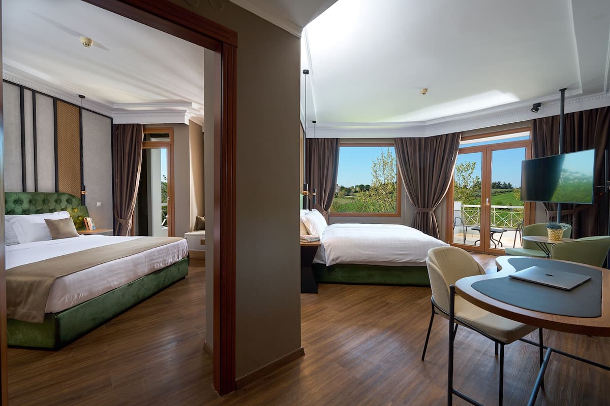 A hotel room with two beds, a dining area with a table and chairs, and a large window offering a view of greenery.
