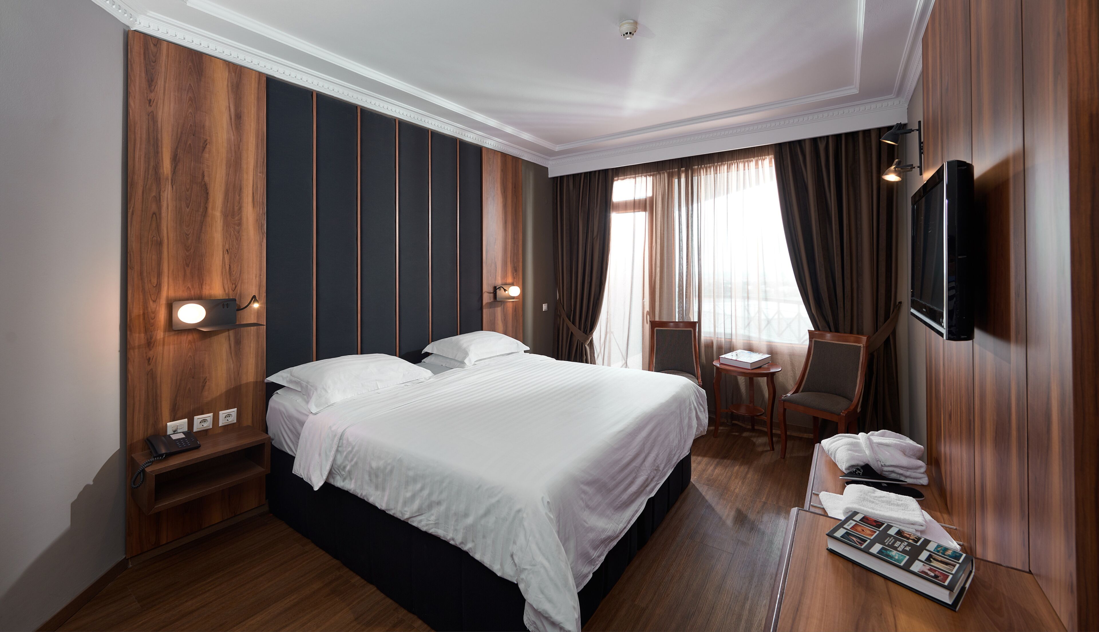 A hotel room with a large bed, a desk with a chair, a TV mounted on the wall, and a window with curtains.