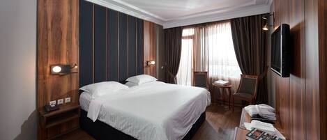 Una habitación de hotel con una cama grande, un escritorio con silla, un televisor montado en la pared y una ventana con cortinas.