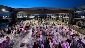 A rooftop dining area with multiple tables and chairs, large windows, and chandeliers.