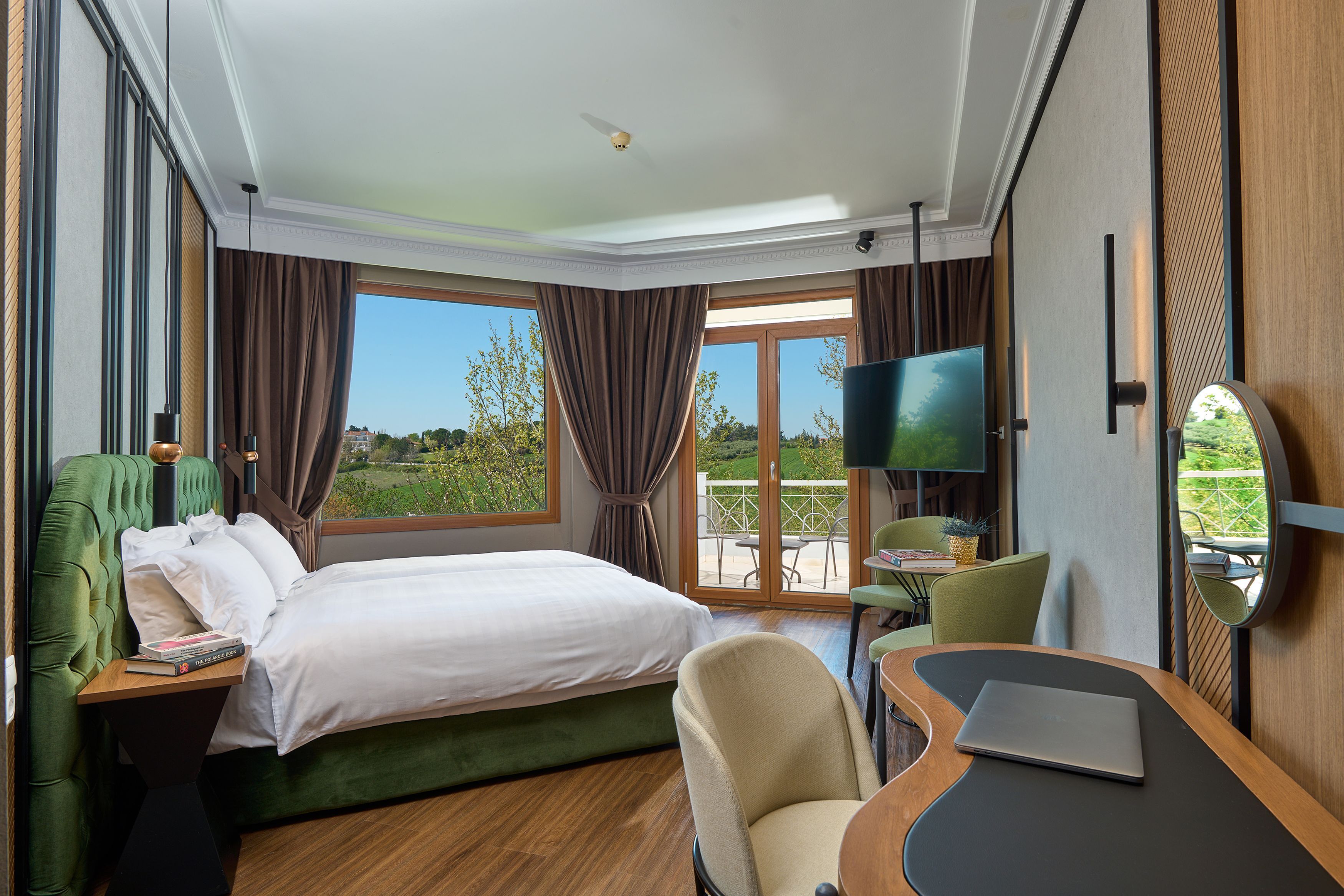 A hotel room with a bed, a desk with a laptop, a chair, a TV, and a balcony with a view of greenery.