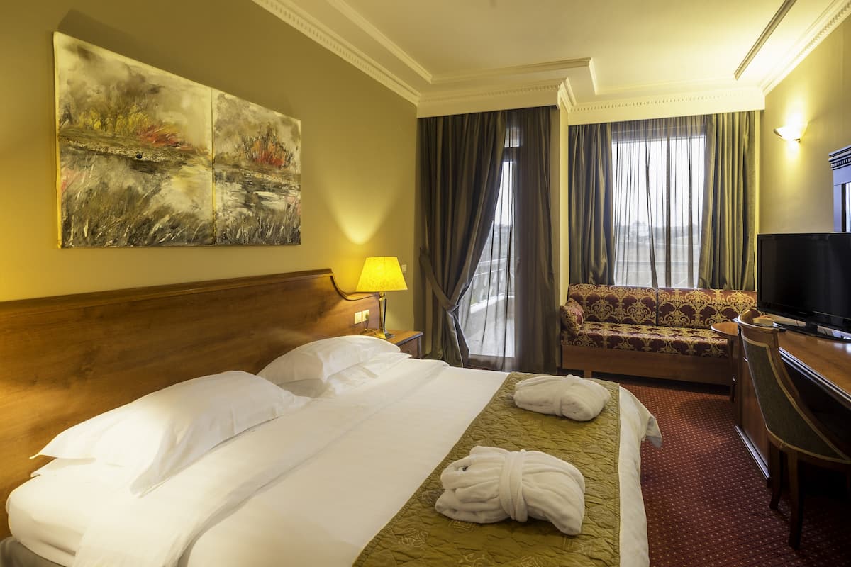 A hotel room with a bed, a sofa, a TV, and a painting on the wall.