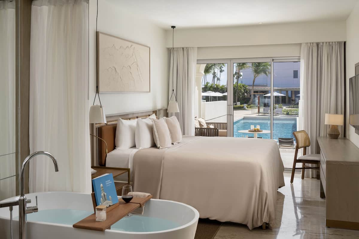 A hotel room with a large bed, a bathtub, a desk, and a view of the pool and palm trees.