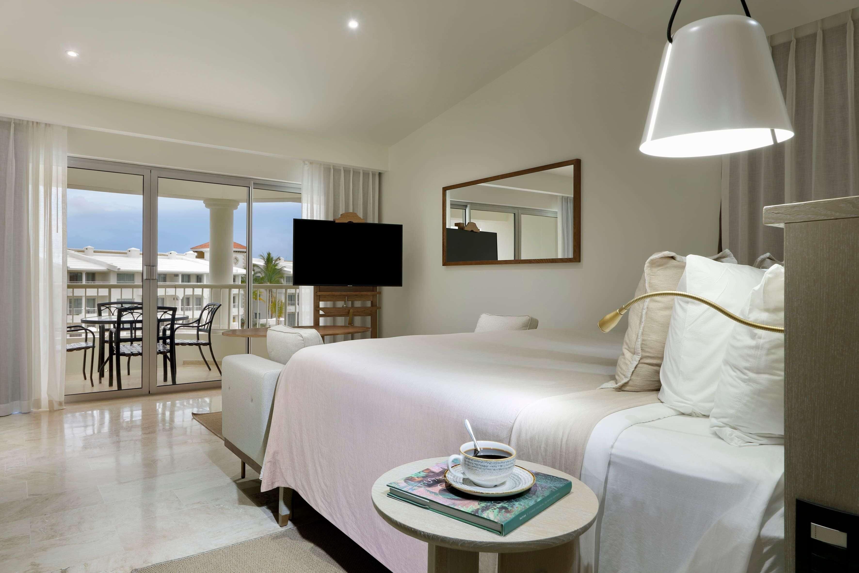 A hotel room with a bed, a television, a round table with a cup and book, and a balcony with chairs.