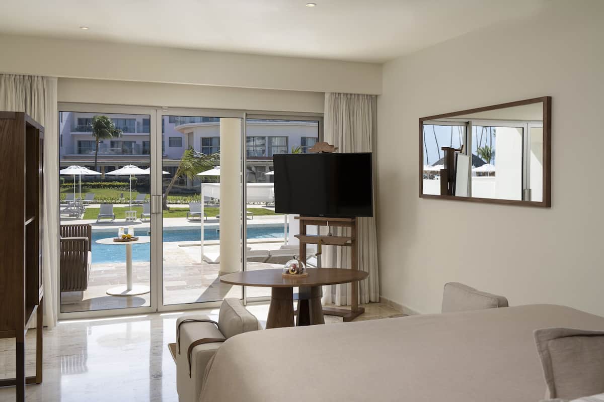 A hotel room with a bed, a TV, a table with a decorative item, and a view of a pool and outdoor seating area.
