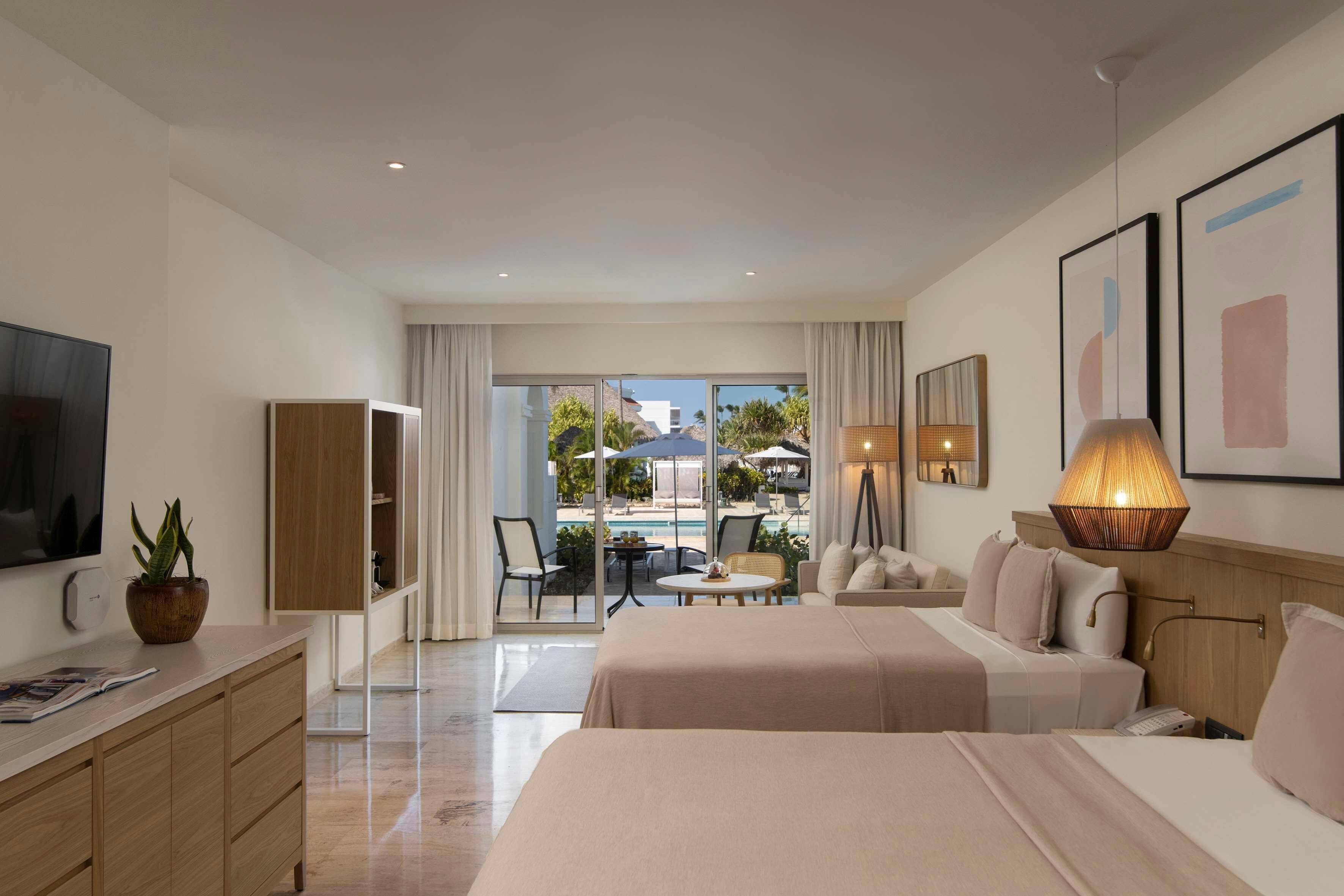 A hotel room with a bed, a desk, a TV, a plant, and a view of the pool.