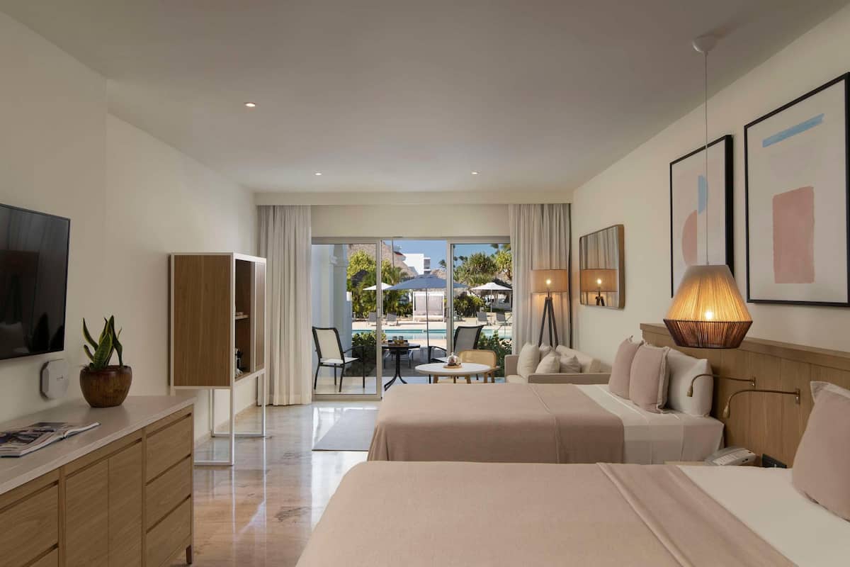 A hotel room with a bed, a desk, a TV, a plant, and a view of the pool.