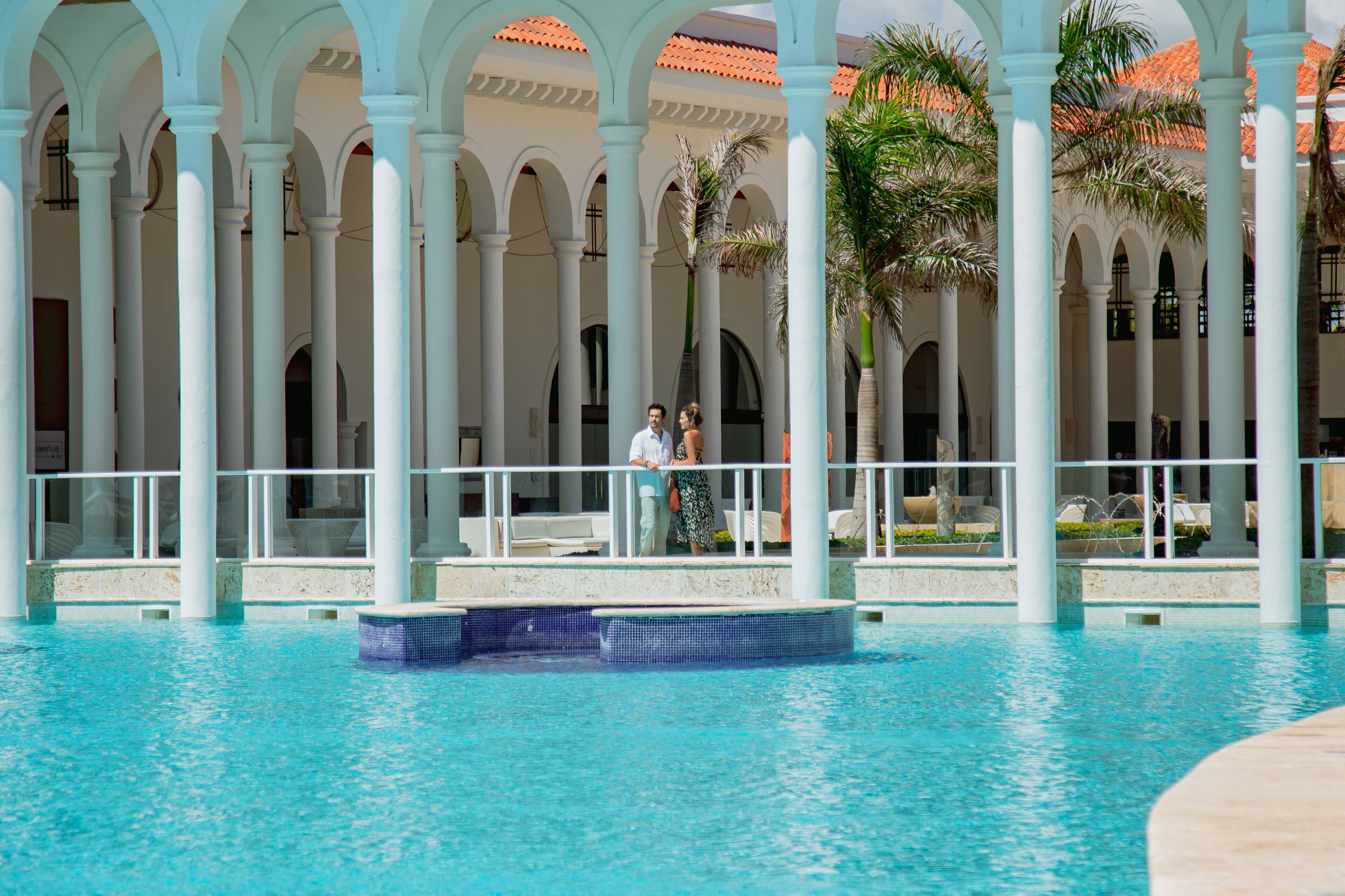 A pool area with a tiled floor, white columns, and palm trees.