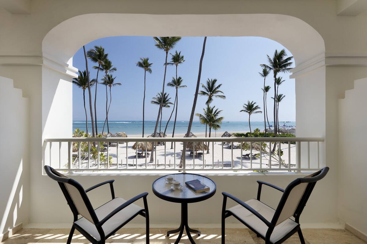 A balcony with two chairs and a table, overlooking a beach with palm trees and the ocean.