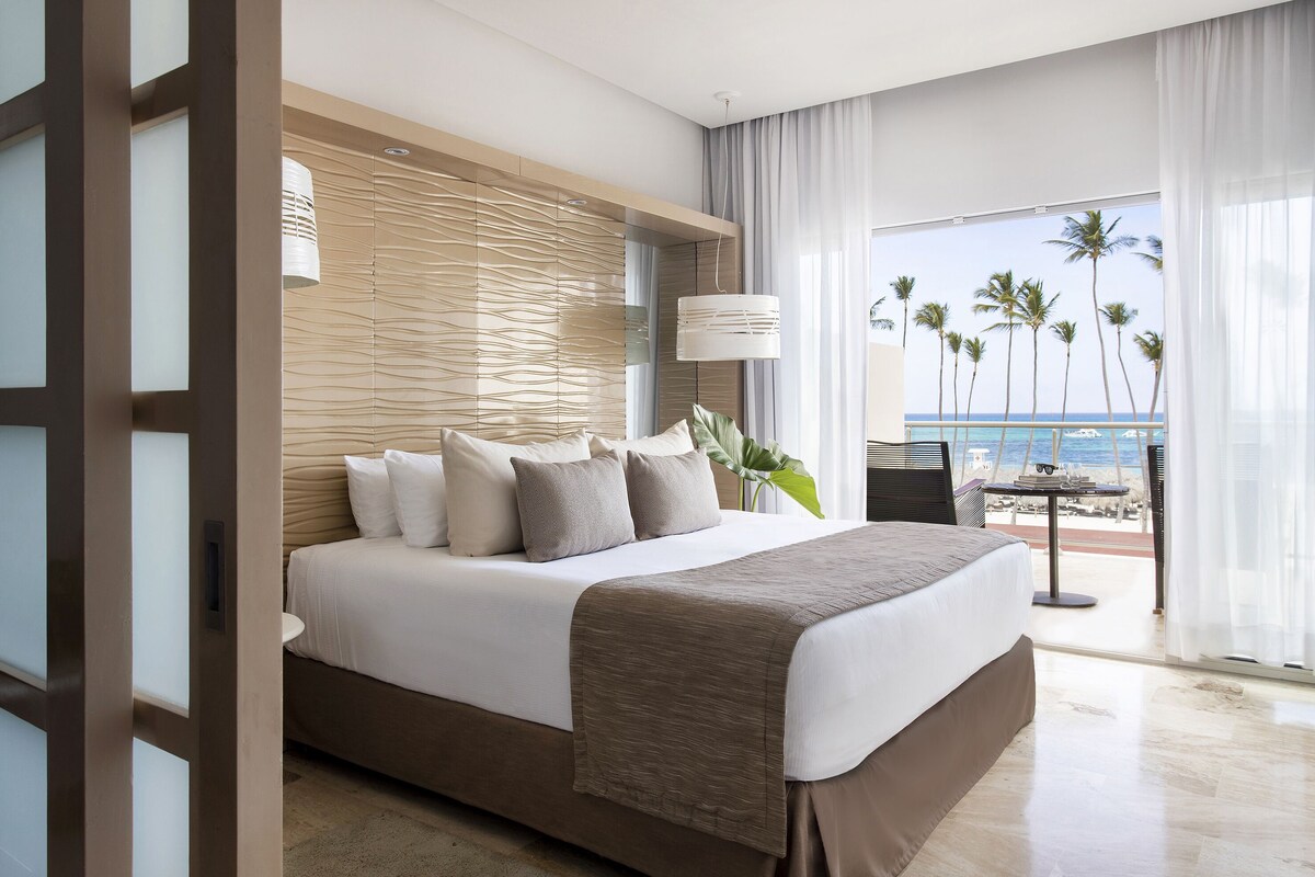 Habitación de hotel con una cama grande, vista a la playa y balcón con mesa y sillas.