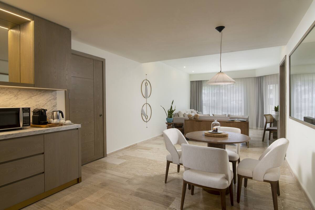 A modern kitchen with a built-in microwave and coffee maker, a dining area with a round table and four chairs, and a large window with sheer curtains.
