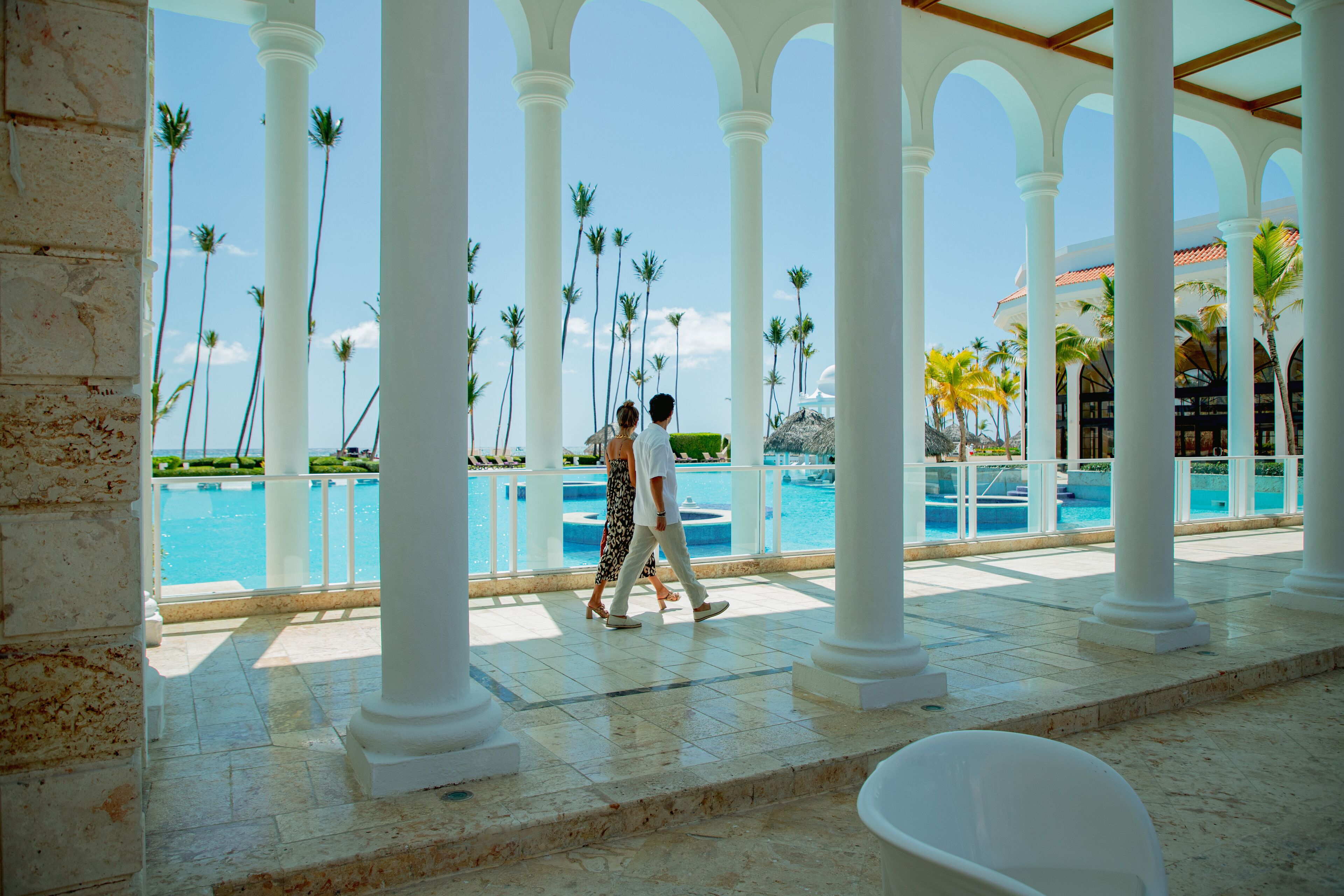 Un espacio al aire libre cubierto con piscina, columnas blancas y palmeras.