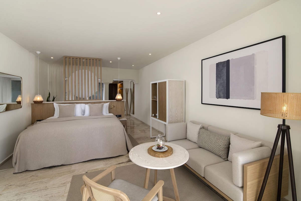 A modern hotel room with a large bed, a sofa, a small round table, and a floor lamp.
