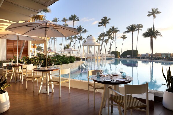 A poolside area with tables and chairs, palm trees, and a clear view of the pool.