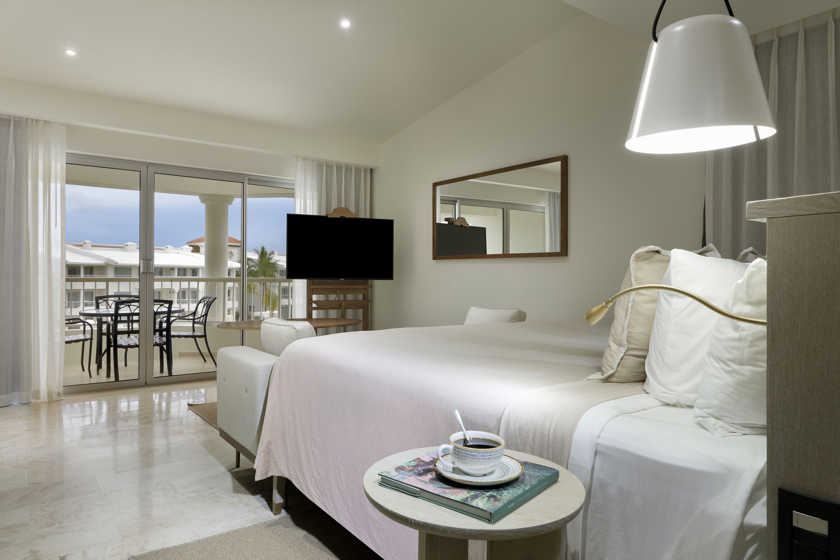 A hotel room with a large bed, a television, a dining area with chairs, and a view through the glass doors.