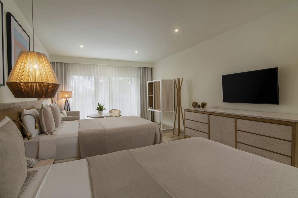 Habitación de hotel con dos camas, un televisor montado en la pared, un escritorio con lámpara y un ventanal con cortinas.