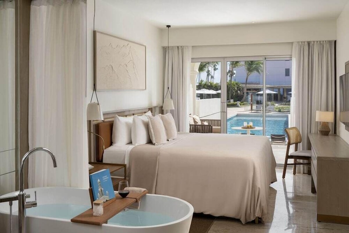 A hotel room with a large bed, a bathtub, a desk, and a view of a pool and palm trees.