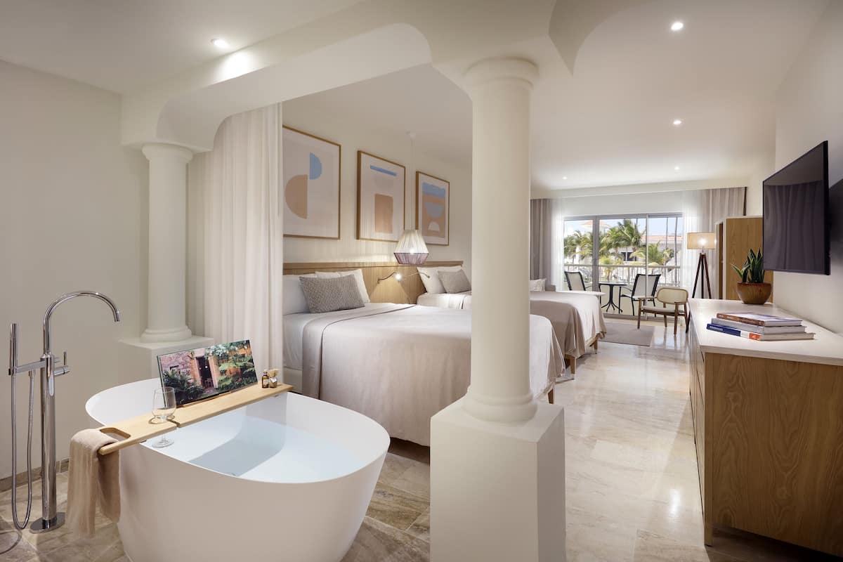 A modern hotel room with a large bathtub, a desk with books, and a view of the outdoors.