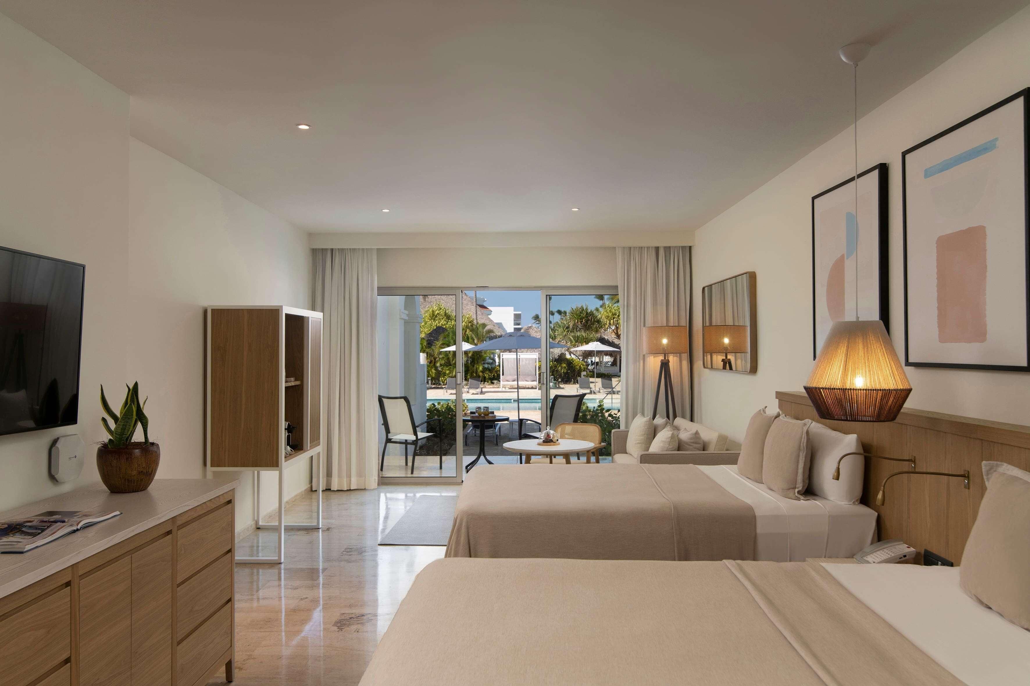 A hotel room with a bed, bedside table, lamp, TV, and a view of the pool and outdoor area.