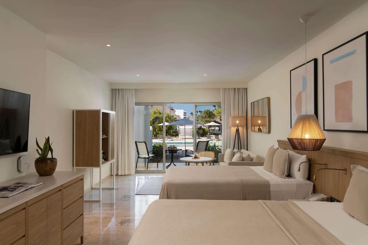 A hotel room with a bed, bedside table, lamp, TV, and a view of the pool and outdoor area.