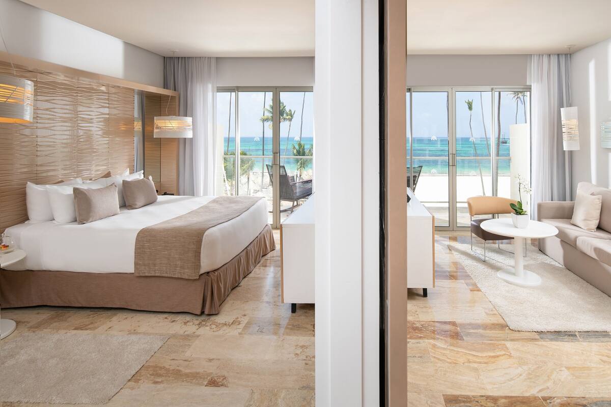 A modern hotel room with a bed, a sofa, a small table, and a view of the beach.