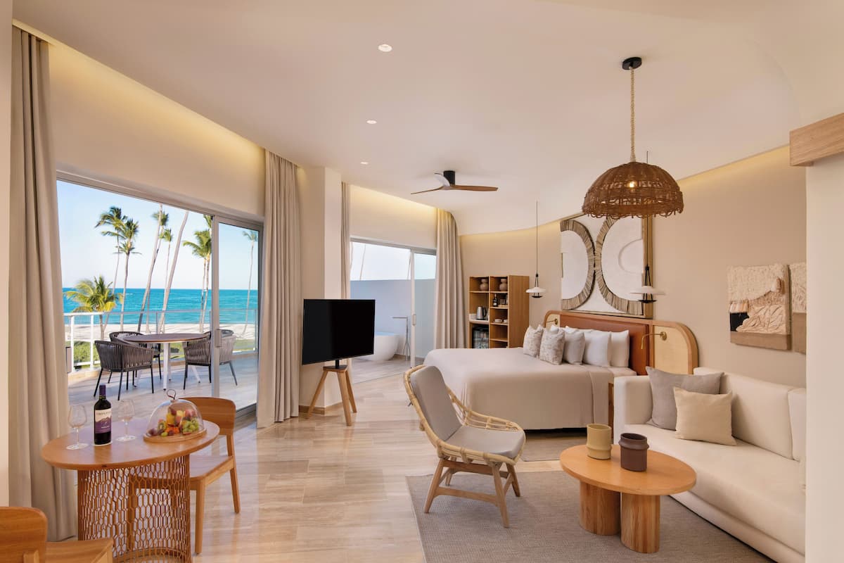 A modern hotel room with a balcony, a bed, a sofa, a dining area, and a TV.