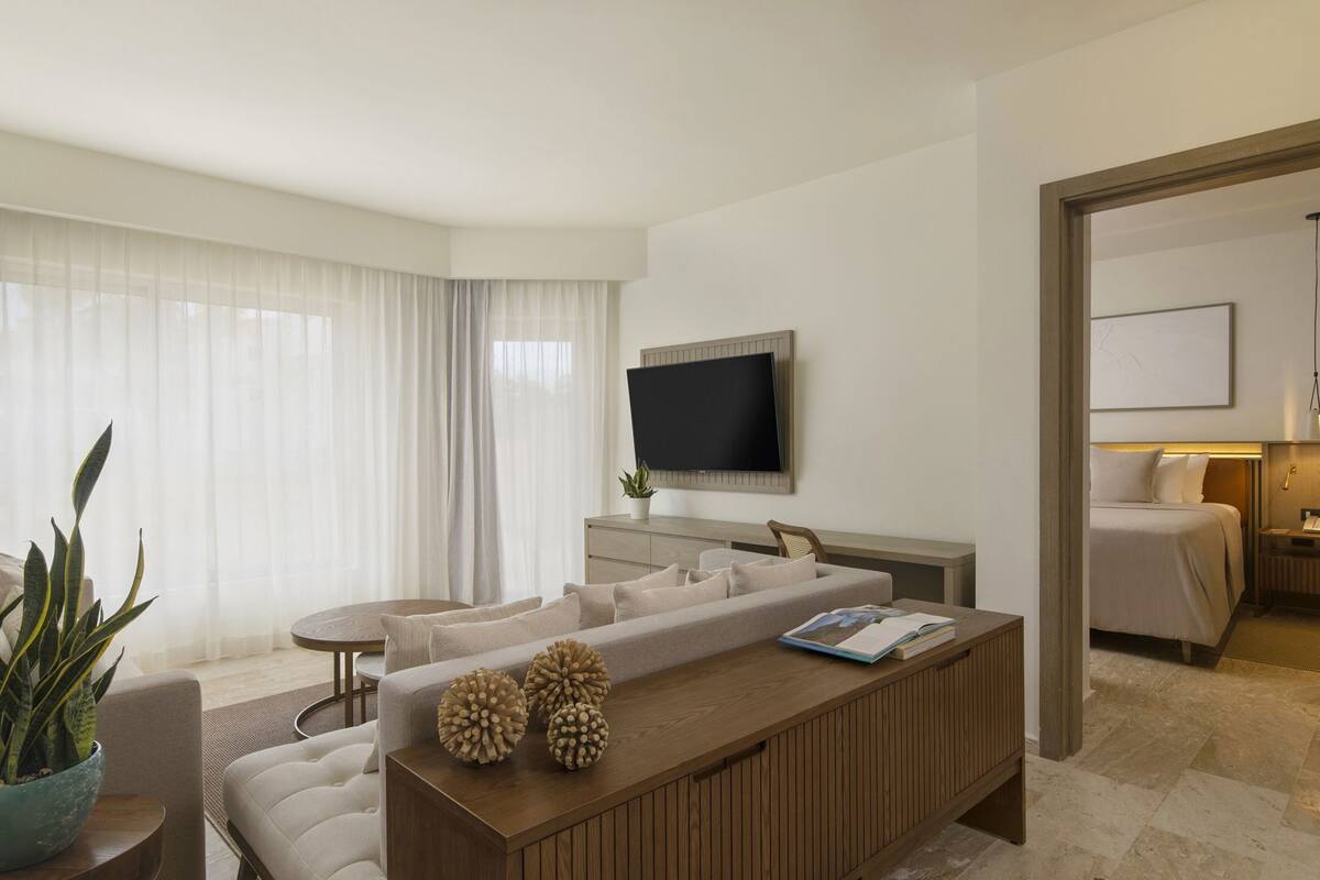 A modern hotel room with a sofa, a wooden coffee table, a TV, and a bed.