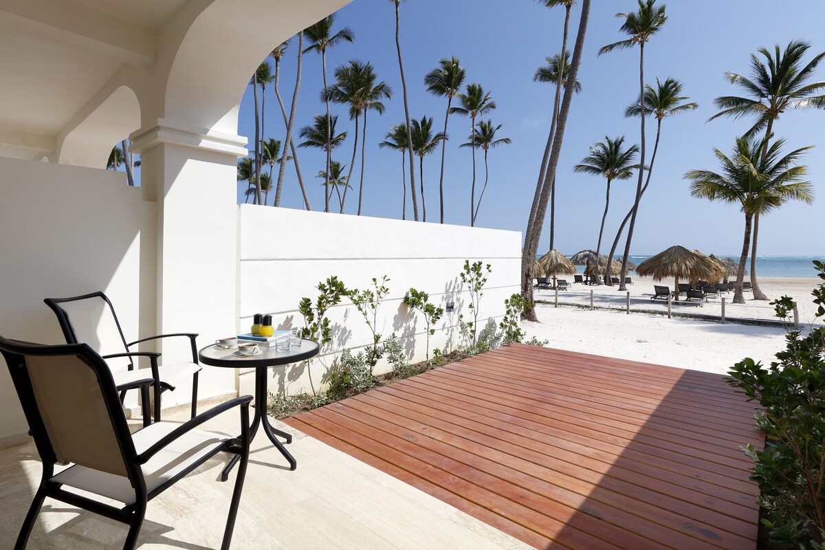 Una terraza de madera con una mesa y sillas, con vistas a una playa con palmeras y una estructura de techo de paja.