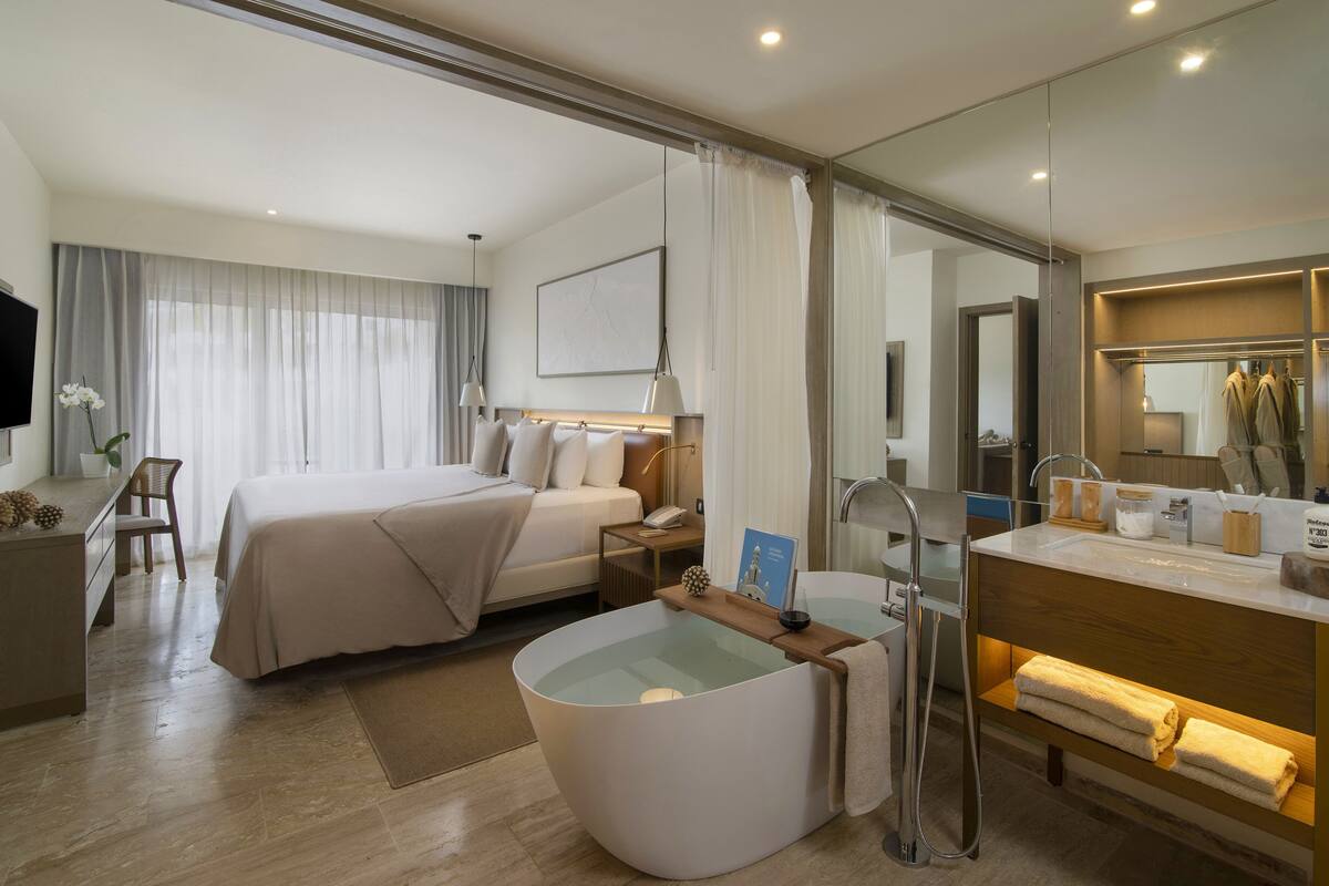A modern hotel room with a large bed, a bathtub, a vanity with a mirror, and a desk with a chair.