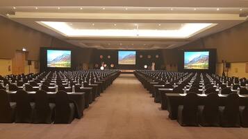 Un salón de conferencias con filas de mesas y sillas, pantallas grandes al frente y piso alfombrado.