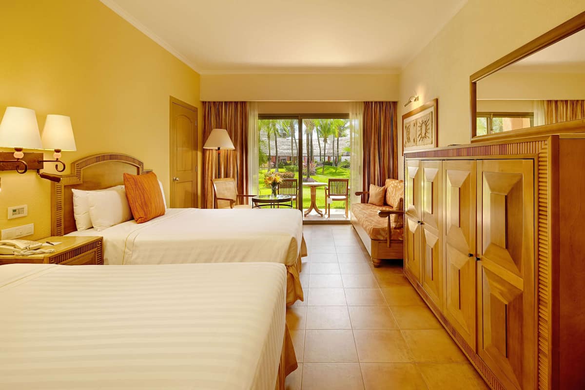 A hotel room with two beds, a desk with a telephone, a chair, a table with a vase of flowers, and a view of a garden through a glass door.