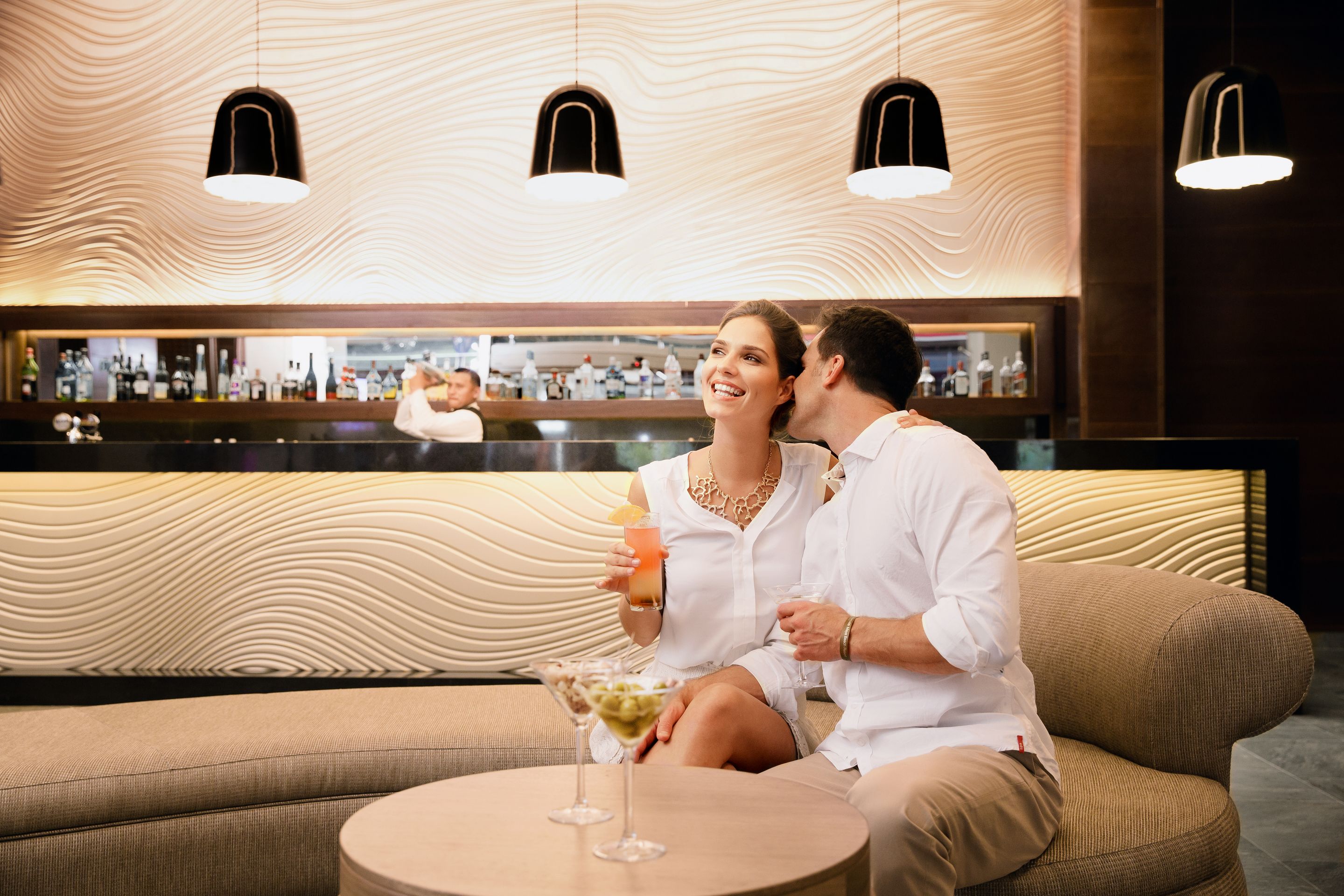 Um casal sentado num sofá numa área de bar moderna, com teto estampado e um bar ao fundo.
