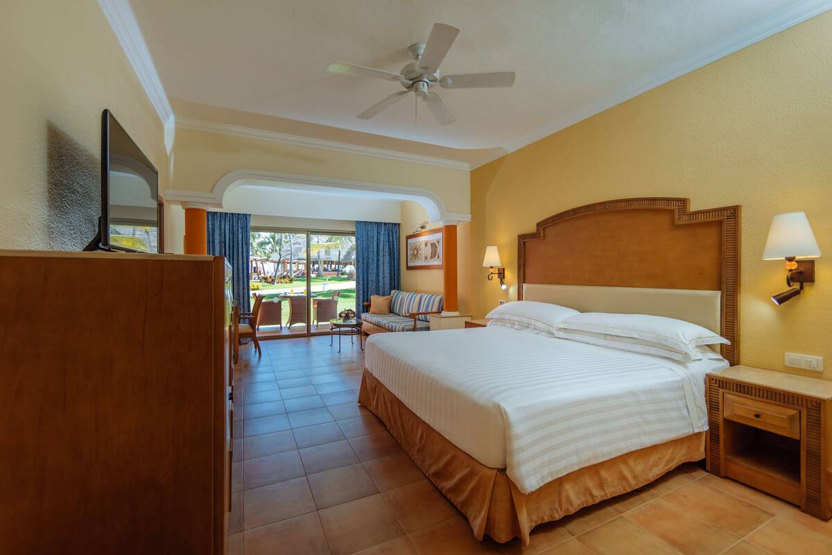 A hotel room with a large bed, a ceiling fan, a TV, a desk, and a view of the outdoor area.