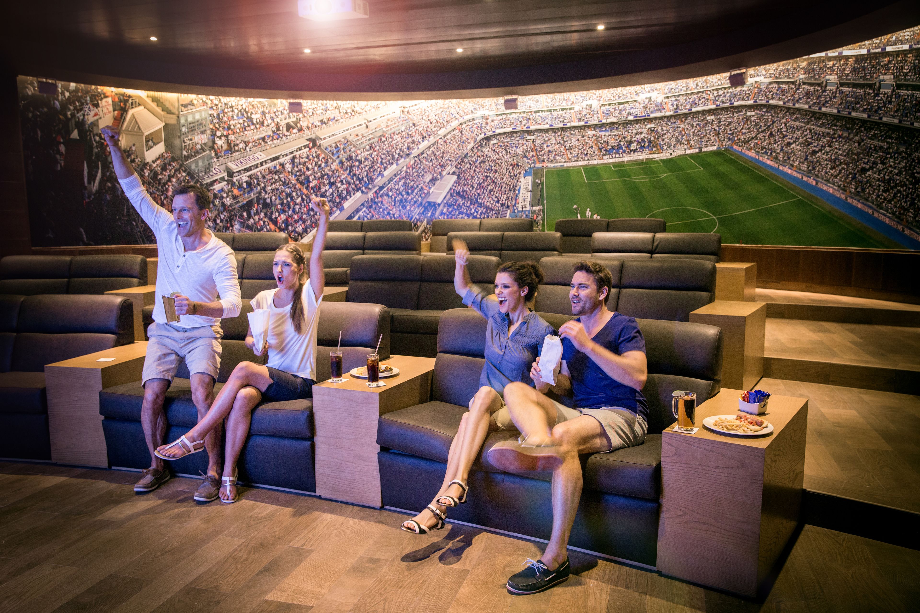 Une salle de cinéma dotée d’un grand écran sur lequel est projetée une image d’un stade. On y voit des rangées de sièges et quelques personnes assises sur un canapé.