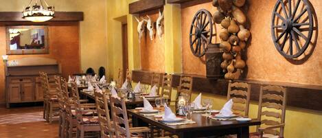 Una larga mesa de comedor dispuesta con sillas, un candelabro y elementos decorativos en la pared, incluyendo calaveras de animales y una rueda.