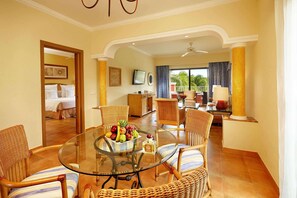 A hotel room with a glass dining table, wicker chairs, a bed with white linens, a television, and a balcony with curtains.