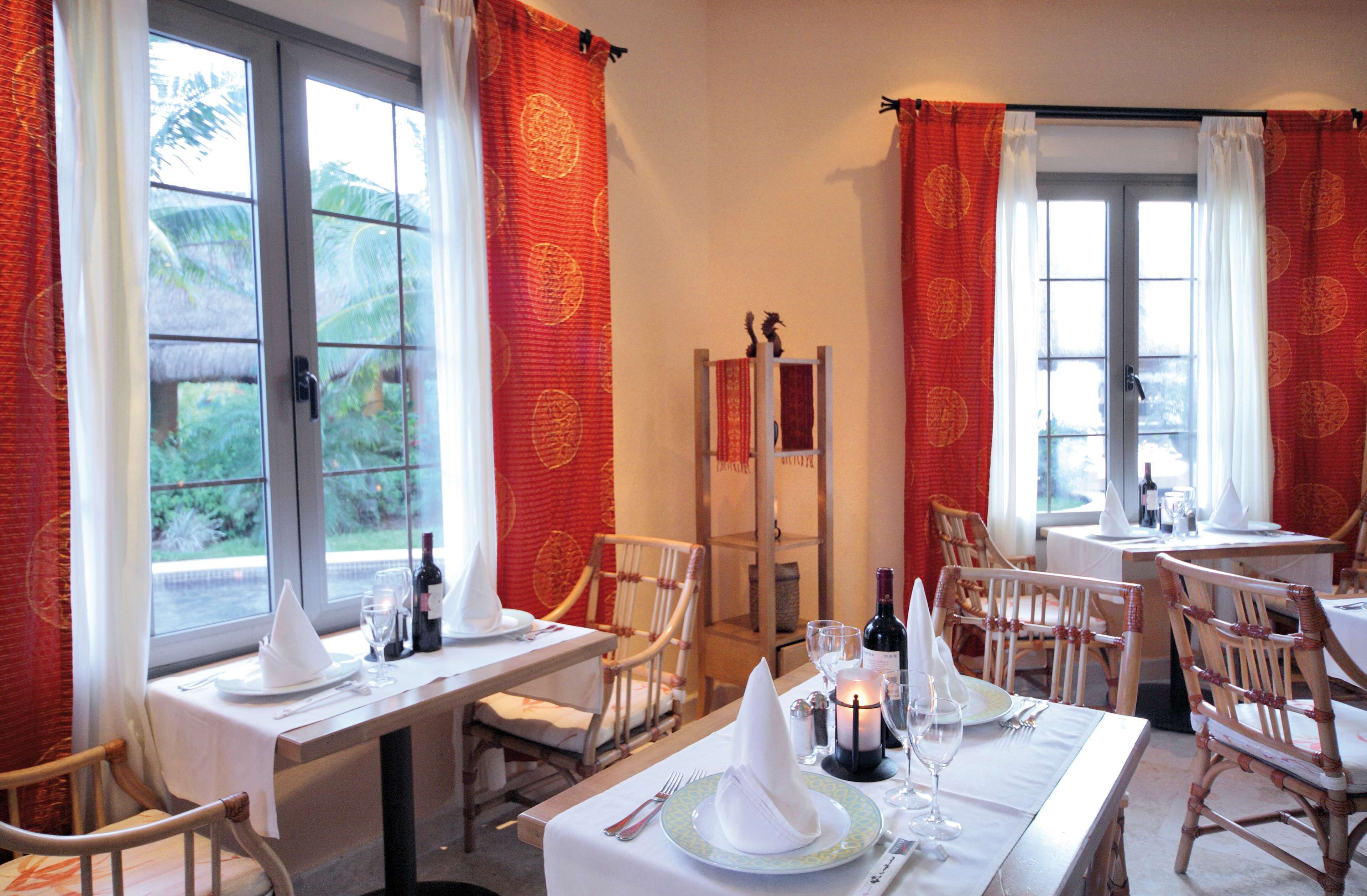 Comedor con mesas dispuestas para dos, sillas y una ventana con cortinas.