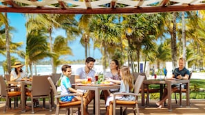 Un grupo de personas cenando en una mesa al aire libre con sillas de mimbre y vistas a palmeras y una playa al fondo.