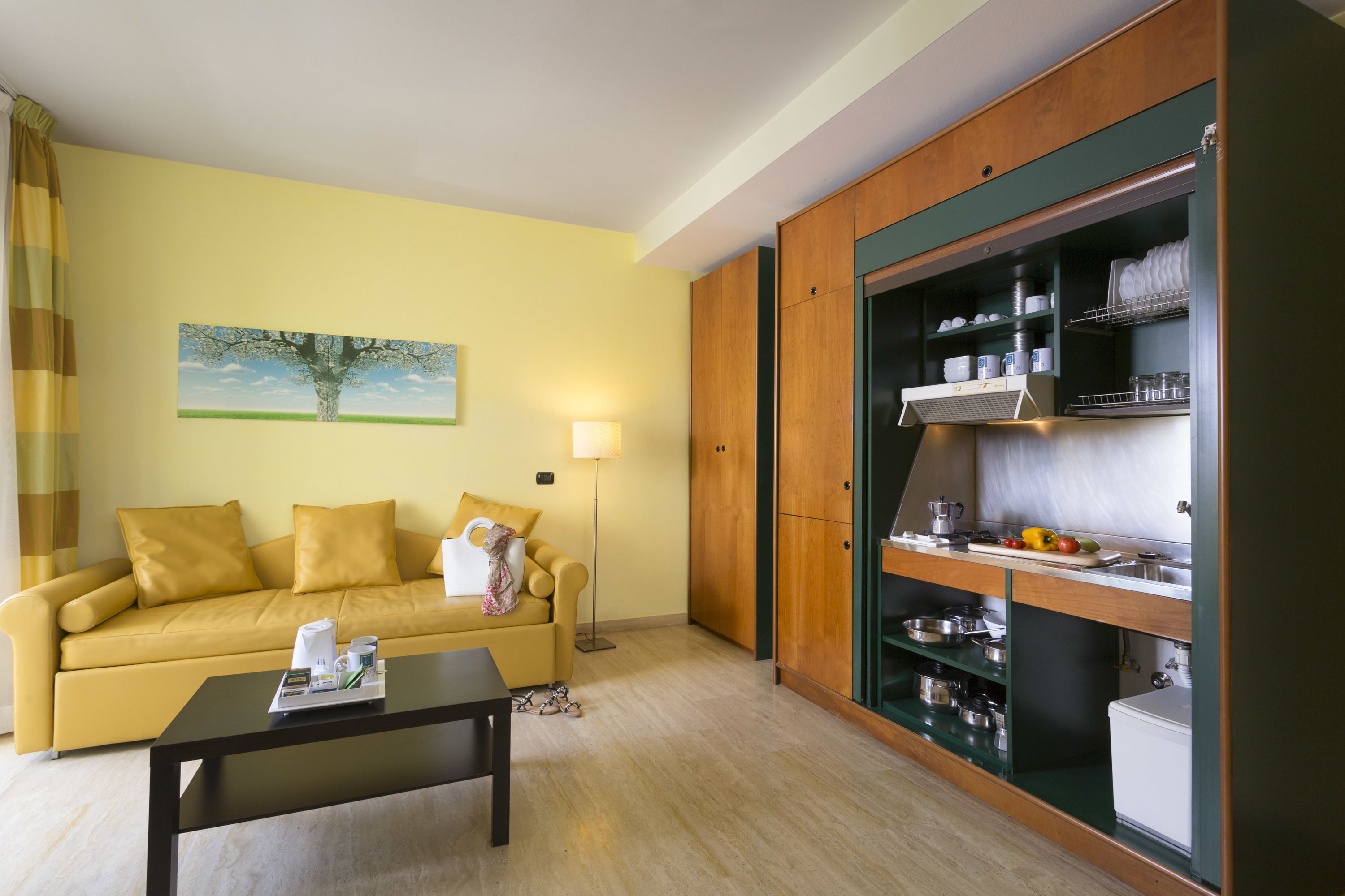 Un sofá amarillo con una mesita de centro, un cuadro de un árbol y un armario de cocina con vajilla.