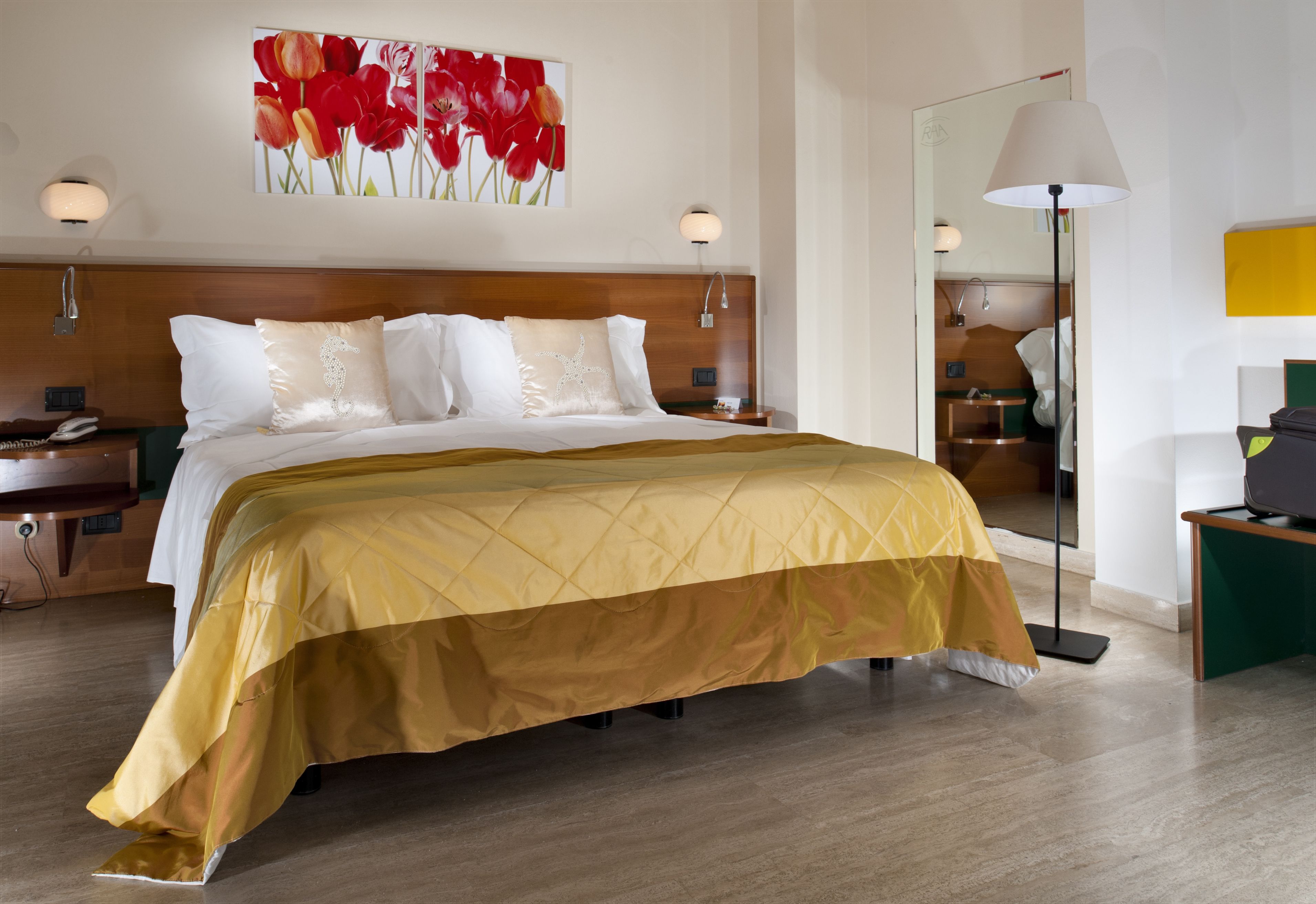 Una camera d'albergo con un letto grande, un comodino, una lampada e un quadro con fiori rossi al muro.